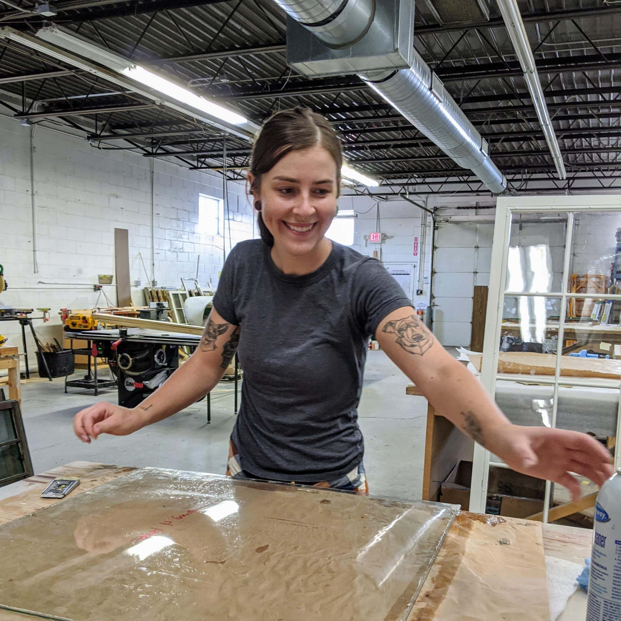 FEMALE WORKER HANDLING A SGLASS PANE SANDWICHED ON A STORE-BOARDED WOODEN TABLE IN A CLOTHED FACTORY ROOM.
