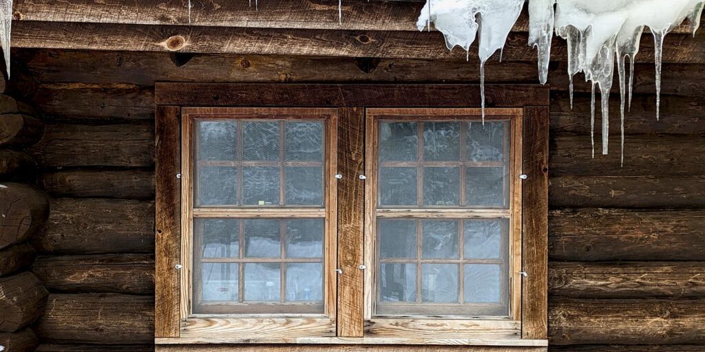Weathered wooden window with ice and snow on log cabin exterior during winter restoration.